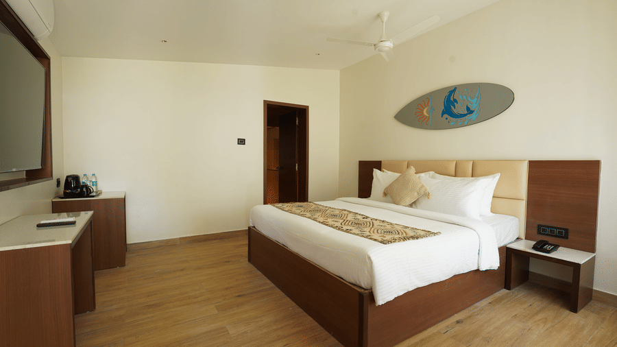 A spacious guest room with a king size bed, light wood floors, a desk area, and a grey surfboard-shaped wall decoration at Trishvam Palolem Beach Resort, Goa.