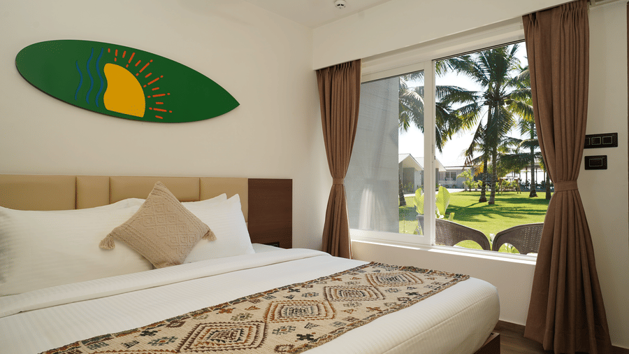 Bedroom with a king size bed, patterned runner, and brown curtains, featuring a window overlooking the palm trees and a green surfboard with a sun logo on the wall at Trishvam Palolem Beach Resort, Goa.
