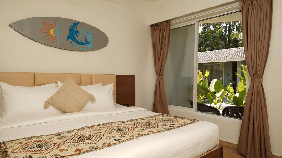 A bed room with patterned runner, large window, surfboard on the wall, and garden view at Trishvam Palolem Beach Resort, Goa.