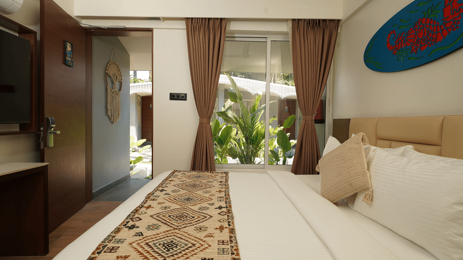 A spacious guest room with a king size bed with tropical garden through an open door and window at Trishvam Palolem Beach Resort, Goa.