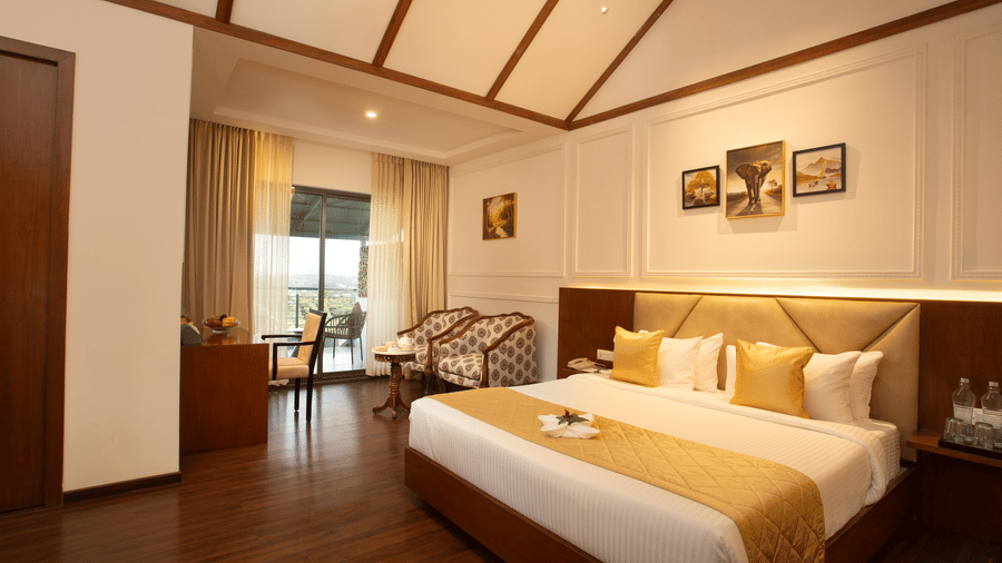 Room at Trishvam Symphony, Krishnagiri featuring large bed with white linens and golden accents, framed artwork, seating area with armchairs, desk with fruit basket, wooden flooring, exposed beam ceiling, and balcony with outdoor seating.