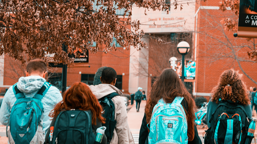 Back view of a group of young people or students wearing rucksacks walking together towards a brick university building.
