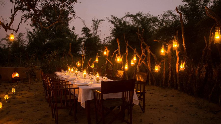 A long dining banquet table set for an outdoor evening meal, beautifully illuminated by hanging oil lanterns along a wooden fence - Umaria Safari Camp
