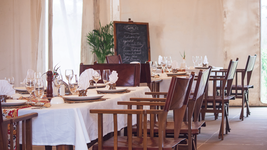 Indoor formal dining arrangement with multiple tables set for a meal, featuring a chalkboard menu in the background - Umaria Safari Camp
