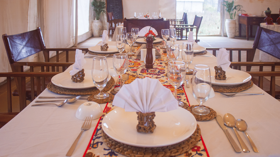A beautifully set long dining table with white table linens, colorful runners, and folded fan napkins inside a bright dining tent - Umaria Safari Camp