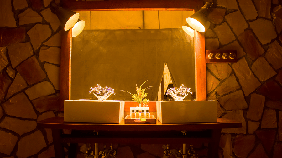 A warmly lit bathroom vanity counter with a large mirror set against a rustic stone accent wall - Umaria Safari Camp
