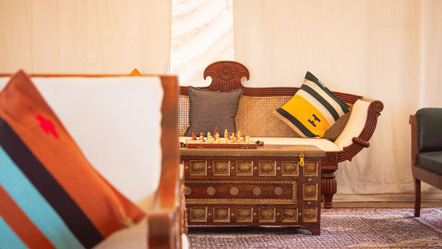Close-up of a rustic-elegant lounge area inside a luxury tent with carved wooden furniture, patterned pillows, and a chess set - Umaria Safari Camp