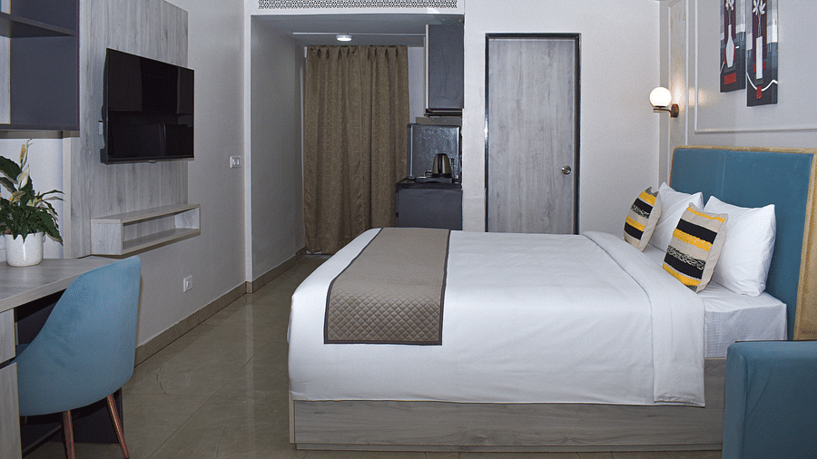 A hotel room with a bed, a desk and chair, a wall-mounted TV, a kitchenette area, a wall cabinet, and partial view of a blue sofa - Urban Suites by BluSalzz, Jaipur