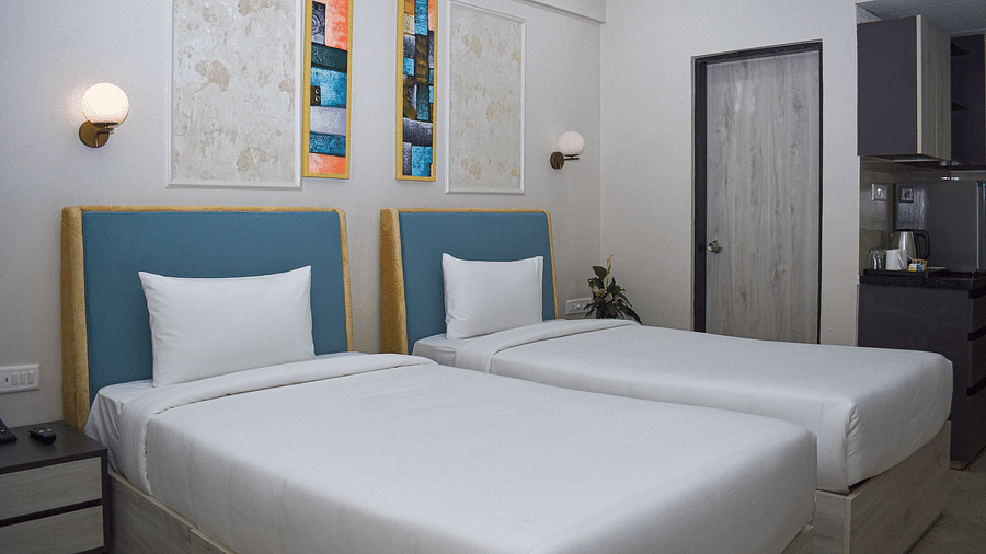 A hotel room with two twin beds, wall-mounted light fixtures, framed artwork, and a partial view of a kitchenette - Urban Suites by BluSalzz, Jaipur