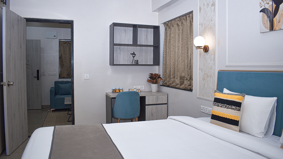 A hotel bedroom with a bed, a desk and chair, a wall cabinet, a window with curtains, and a doorway leading to another room - Urban Suites by BluSalzz, Jaipur