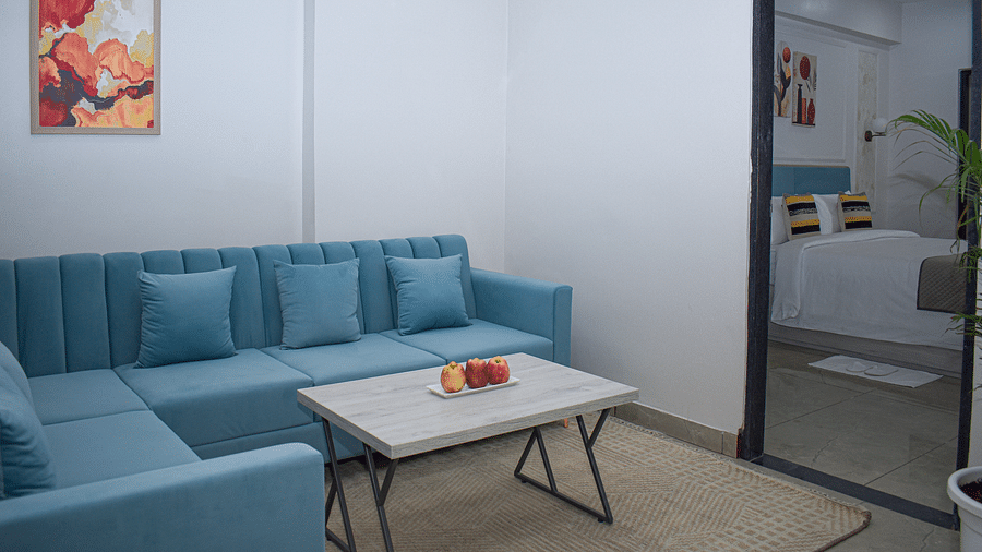 A hotel room living area with a blue sofa, a light wood coffee table with fruit, a rug, and a view into the bedroom - Urban Suites by BluSalzz, Jaipur