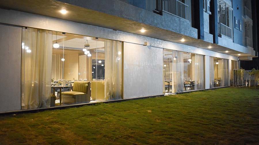 Exterior view of a building with a glass-walled restaurant, sheer curtains, hanging lights, and a grassy area in front - Urban Suites by BluSalzz, Jaipur