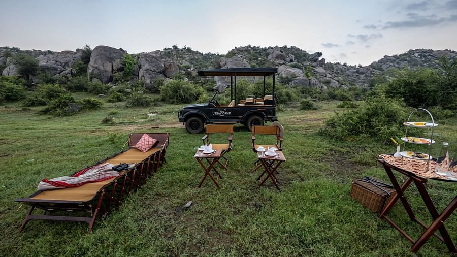 An image of an outdoor seating arrangement with a jeep parked nearby amidst greenery and the backdrop of green mountains - Utsav Camp Sariska