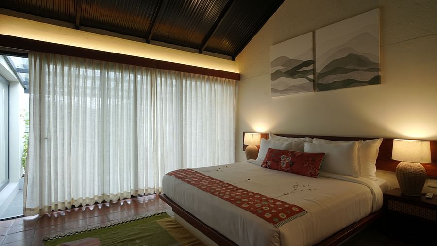 A deluxe room at Vaasta Vagamon featuring a large bed with decorative throw pillows and a patterned runner, and long drapes.