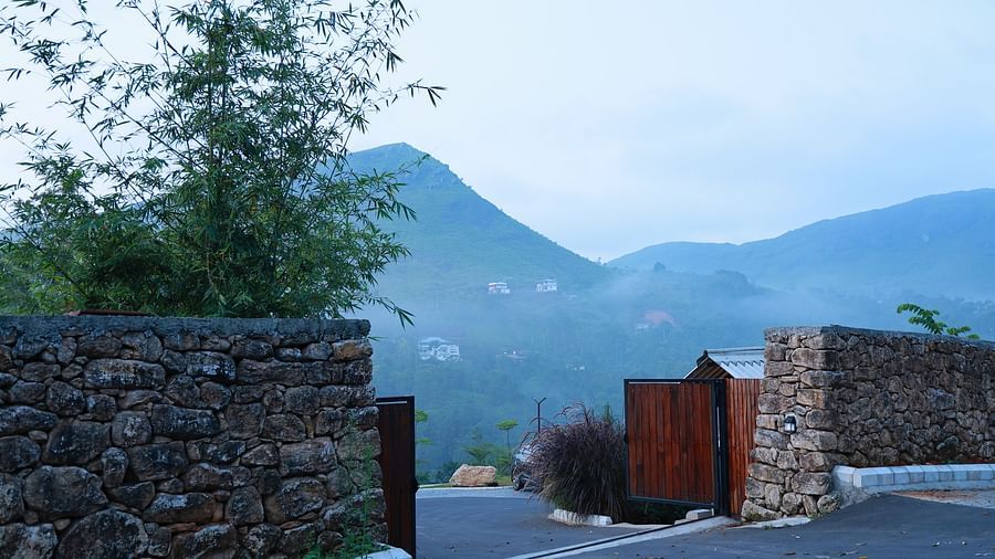 A stone wall entrance with an open wooden gate at Vaasta Vagamon, revealing a winding road and distant mist-shrouded hills.
