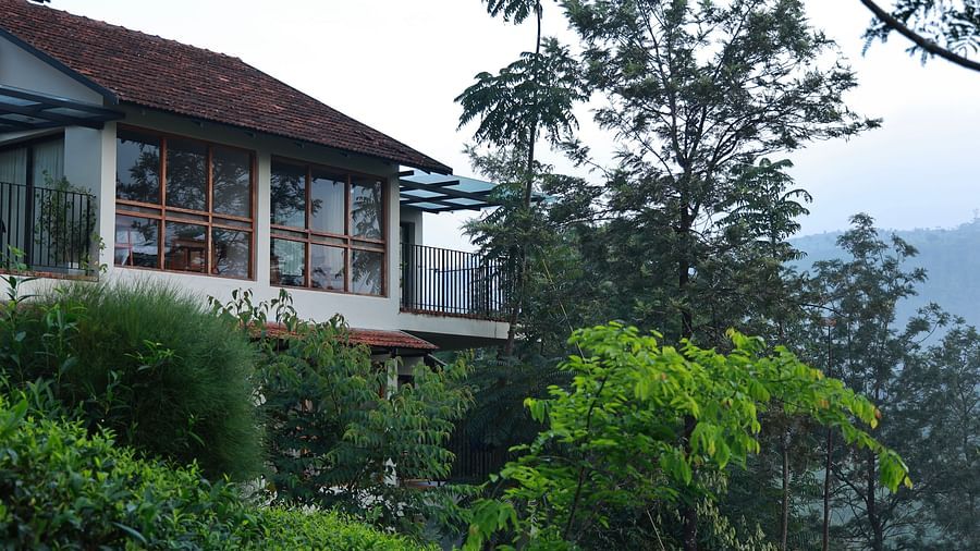  A building at Vaastha Vagamon, nestled amidst abundant foliage, with large windows overlooking a verdant landscape.