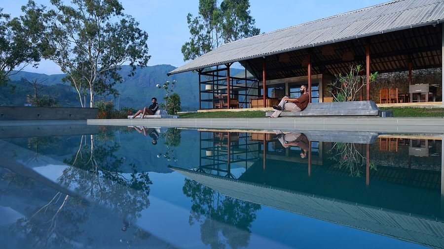 Two people relax by an infinity pool at Vaasta Vagamon, overlooking a serene landscape with rolling hills and trees.