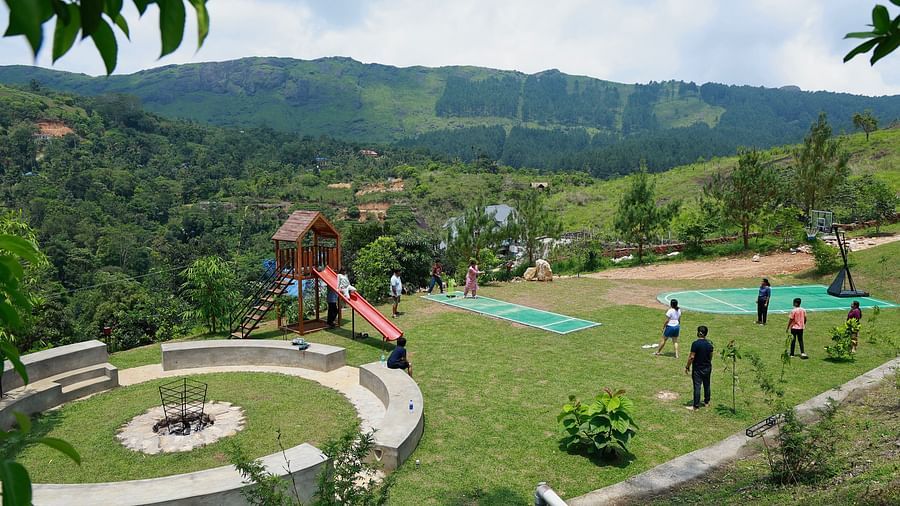  A vibrant outdoor recreational area at Vaastha Vagamon with a playground, sports court, and fire pit, surrounded by lush hills.