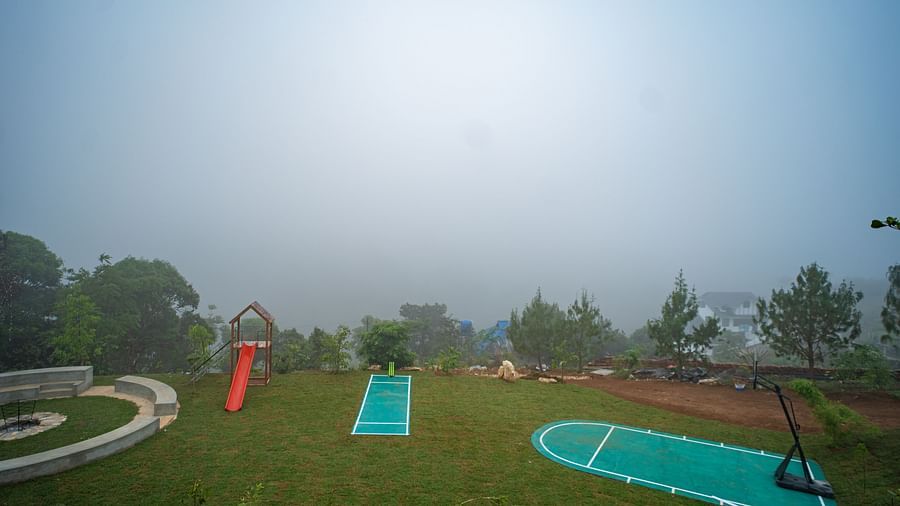  A vast outdoor recreational area at Vaastha Vagamon, featuring a playground, sports courts, and a fire pit, all enveloped in mist.