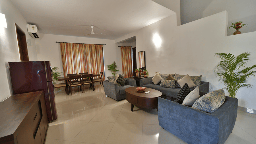 A spacious living area of the Aqua Bungalow at Vedic Village Spa Resort featuring blue sofas wooden furniture indoor plants tiled flooring and warm natural light from large windows.
