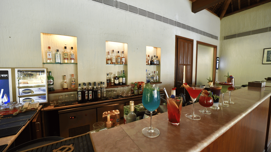 A bar counter at Vedic Village Spa Resort featuring colorful cocktail glasses liquor bottles a display refrigerator and warm ambient lighting in a thatched roof setting.