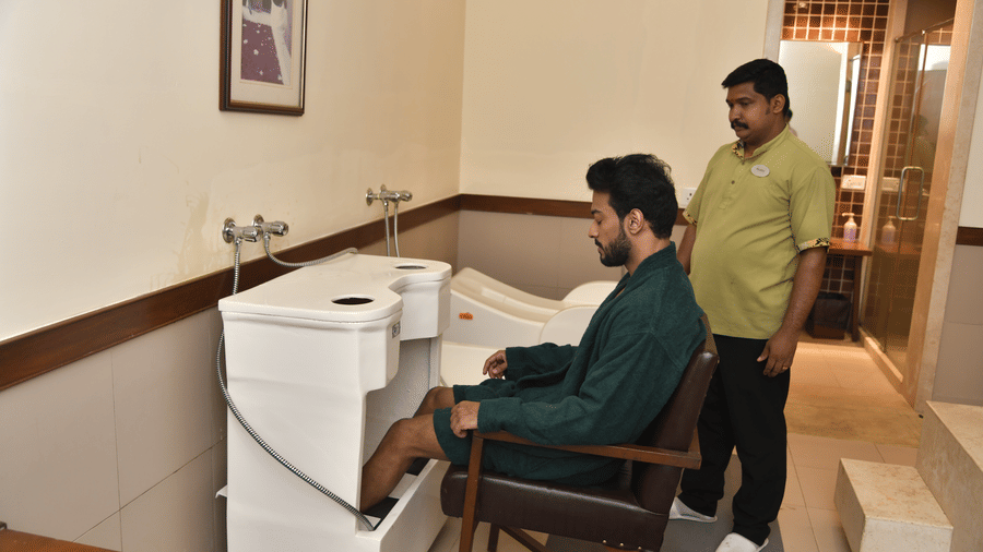 A therapist assisting a male guest during a colon hydrotherapy session at Vedic Village Spa Resort in a clean treatment room with specialized equipment and white tiled walls.