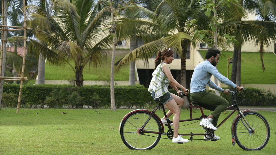A couple cycling together on a lush green lawn at Vedic Village Spa Resort surrounded by tall palm trees and open green spaces in a bright outdoor setting.