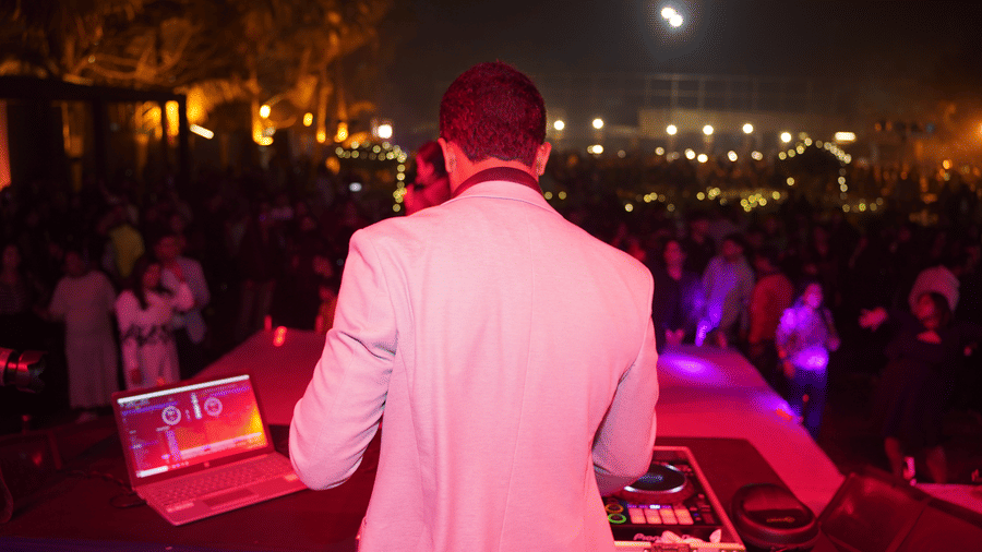 A DJ performing at an outdoor night event at Vedic Village Spa Resort with a large crowd gathered in the background under warm ambient and purple stage lighting.