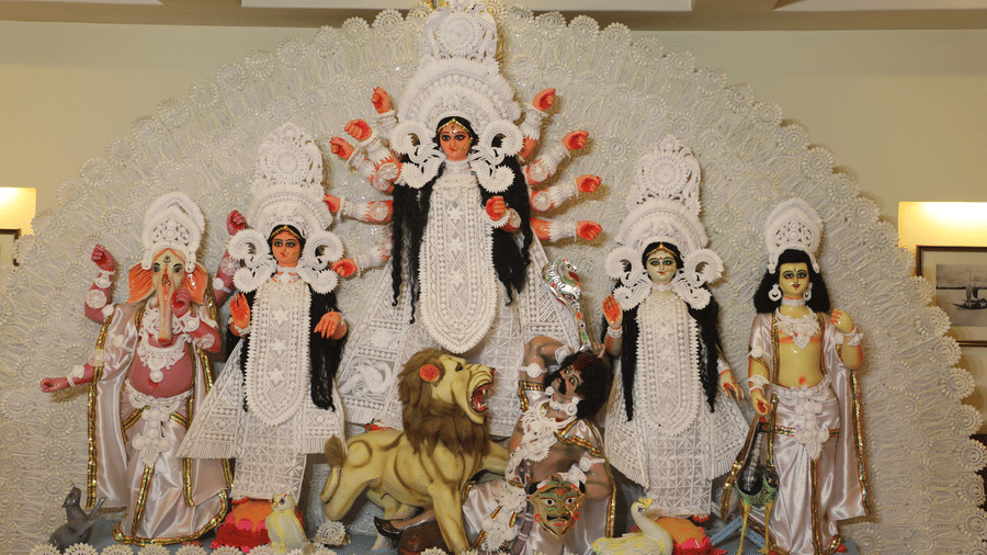 A full view of the Durga Puja idol display at Vedic Village Spa Resort featuring the complete ensemble of deities decorated with white floral arrangements in an indoor pandal setting.