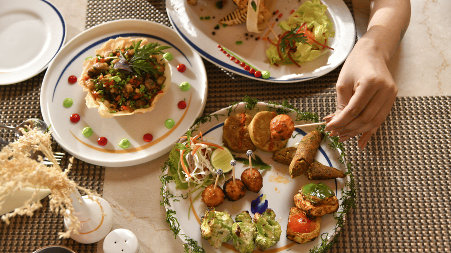 An overhead view of a server placing a platter of assorted dishes on a table at Vedic Village Spa Resort with multiple food items and condiments arranged on a woven surface.