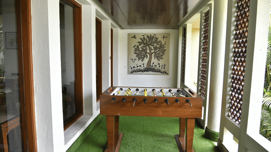 An indoor games room at Vedic Village Spa Resort featuring a foosball table green artificial turf flooring large windows and a framed tree artwork on the white wall.