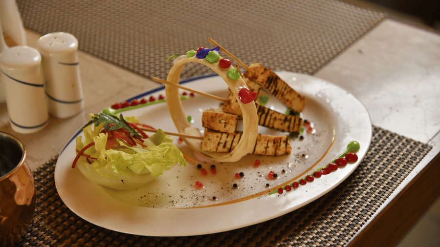 A gourmet food platter at Vedic Village Spa Resort featuring artistically arranged appetizers with garnish and condiments served on a white plate with a wooden surface beneath.