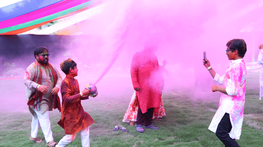 A group of people celebrating Holi outdoors at Vedic Village Spa Resort with vibrant pink and multicolored powder filling the air in a festive and joyful setting.