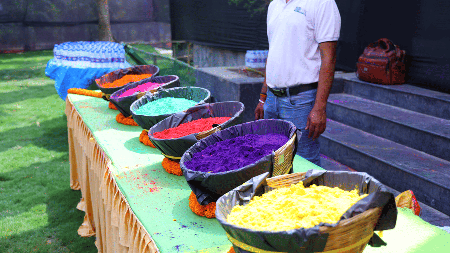 A Holi celebration setup at Vedic Village Spa Resort featuring large bowls filled with colorful powder arranged on a decorated table with a staff member standing alongside.