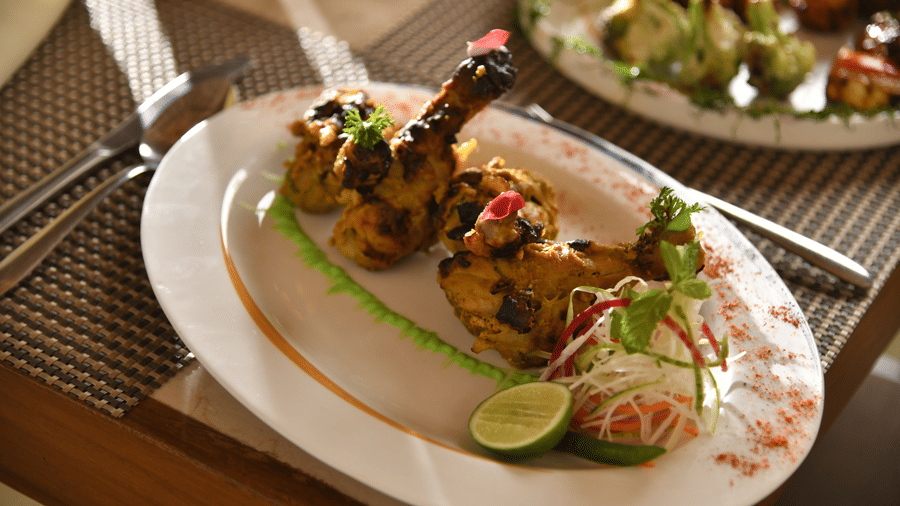 A platter of grilled kebabs served with lime garnish and accompaniments on a white plate at Vedic Village Spa Resort presented on a textured woven table surface.