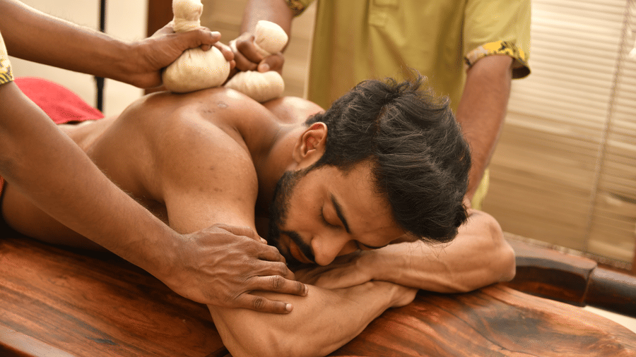 Male guest lying face down on a wooden massage table receiving a kizhi herbal pouch massage treatment by a therapist at Vedic Village Spa Resort.
