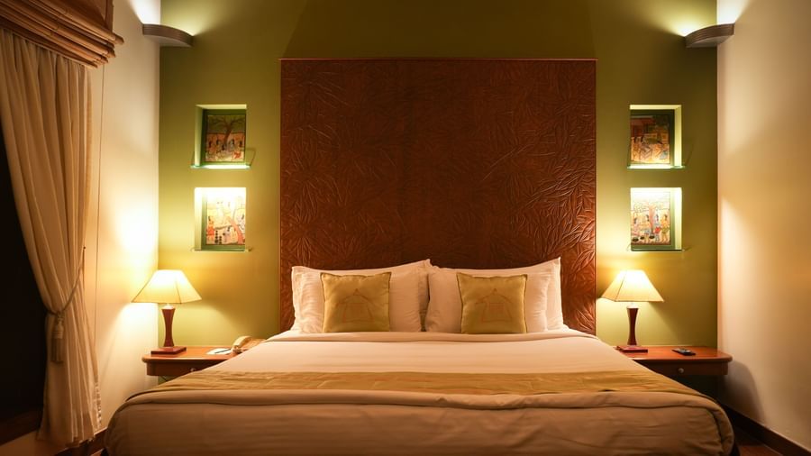Front angle view of the bedroom in the Lake View Villa at Vedic Village Spa Resort showing a neatly made queen size bed and a framed painting under soft lighting.