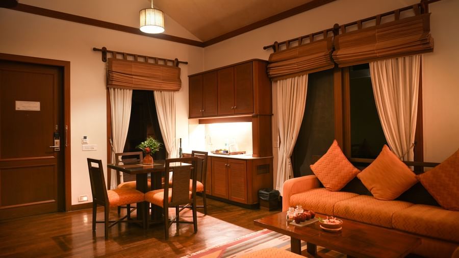 Warmly lit living area in the Lake View Villa at Vedic Village Spa Resort with in-room dining space, carpeted floors, plush couch, and wooden furnishings.