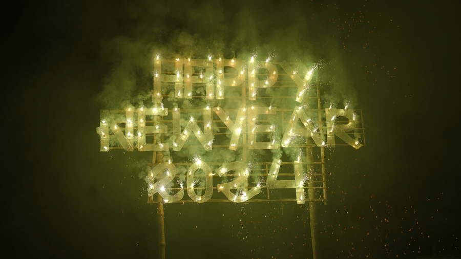 A glowing Happy New Year 2024 light installation displayed at night during a New Year celebration event at Vedic Village Spa Resort against a dark sky background.
