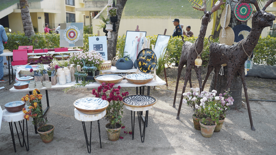 An outdoor decor and art display at Vedic Village Spa Resort featuring decorative deer sculptures round tables floral arrangements potted plants and various artwork under shaded trees.