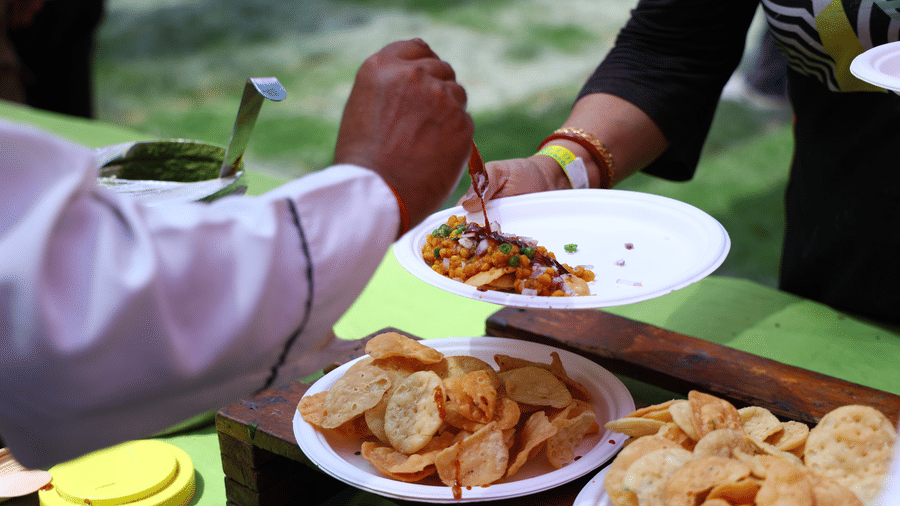 A guest being served traditional snacks on a plate at an outdoor food service event at Vedic Village Spa Resort with green lawns and natural surroundings in the background.