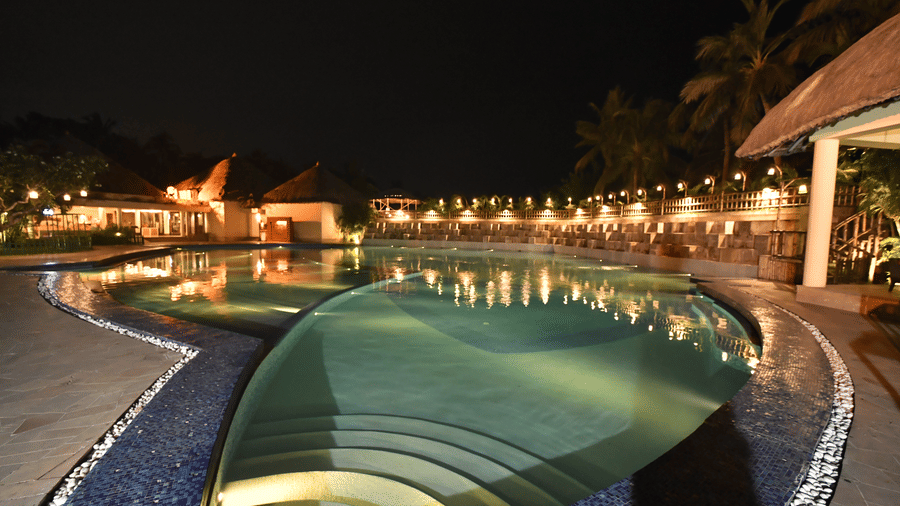 An aerial night view of the swimming pool at Vedic Village Spa Resort featuring a curved pool with warm lighting surrounding resort buildings and tall palm trees under a dark sky.
