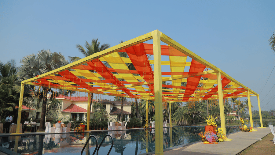 A colorful yellow and orange canopy structure set up beside the swimming pool at Vedic Village Spa Resort with tropical trees and resort buildings visible in the background.