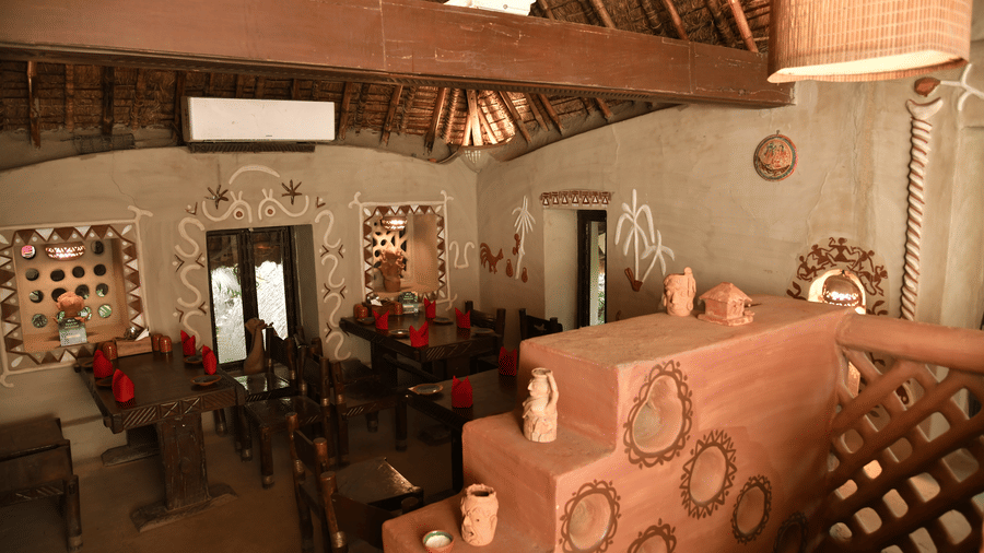 Interior view of a rustic restaurant at Vedic Village Spa Resort featuring terracotta walls thatched roof wooden furniture red table settings and traditional wall art decor.