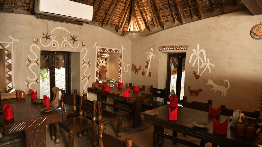 A wide view of the restaurant interior at Vedic Village Spa Resort featuring a high thatched roof red table settings wooden furniture and traditional white wall art on terracotta walls.