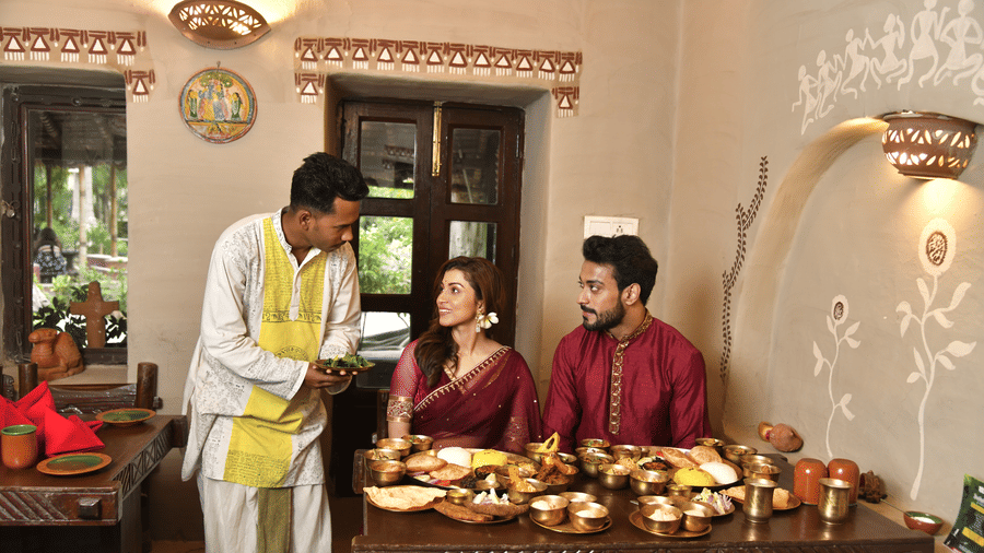 A staff member in white attire serving a couple in traditional outfits at a dining table at Vedic Village Spa Resort with brass utensils and warm ambient lighting in the background.