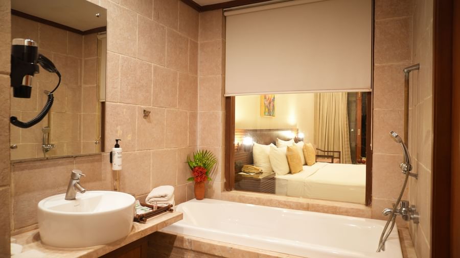 Washroom of the Standard Suite at Vedic Village Spa Resort featuring a sink, wall mirror, and a bathtub set against a glass-panelled wall overlooking the room, with towels and amenities placed beside the sink.