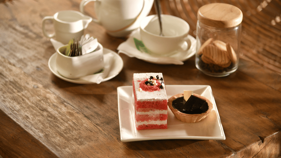 A close-up of a tea and dessert setup at Vedic Village Spa Resort featuring a teacup saucer and a small dessert plate with cake and garnish on a wooden table surface.A close-up of a tea and dessert setup at Vedic Village Spa Resort featuring a teacup sauce