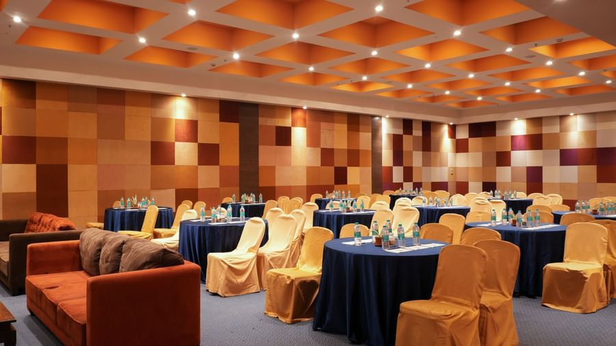 The V-den event hall in Vedic Village Spa Resort, featuring brown cushioned couch, yellow chairs, round tables covered in blue and modern interiors.
