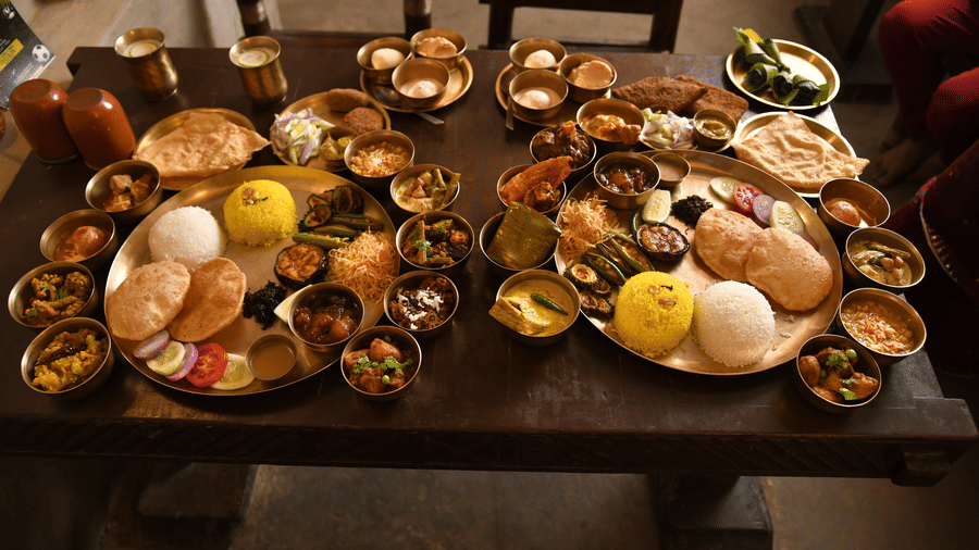 A traditional Bengali thali spread at Vedic Village Spa Resort featuring multiple brass bowls and plates filled with various dishes arranged on a long wooden dining table.